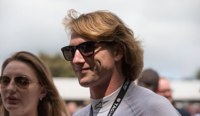 Man with light brown hair wearing dark sunglasses and a lanyard at an outdoor event; smiling slightly, with a woman wearing sunglasses beside him in the foreground.