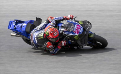 Motorcycle racer in blue Yamaha livery leaning into a sharp turn on a grey racetrack, Monster Energy branding visible on the bike and suit.