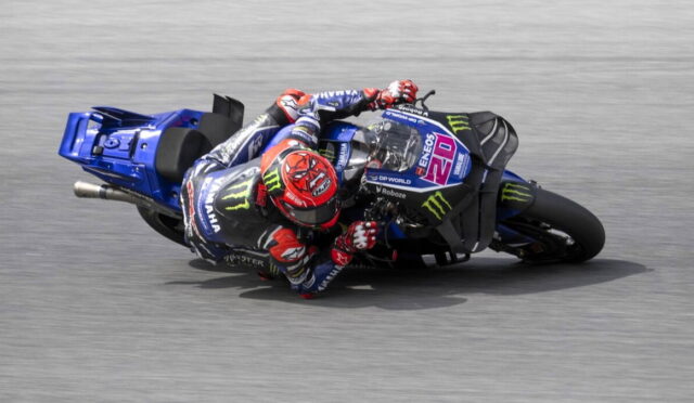 Motorcycle racer in blue Yamaha livery leaning into a sharp turn on a grey racetrack, Monster Energy branding visible on the bike and suit.
