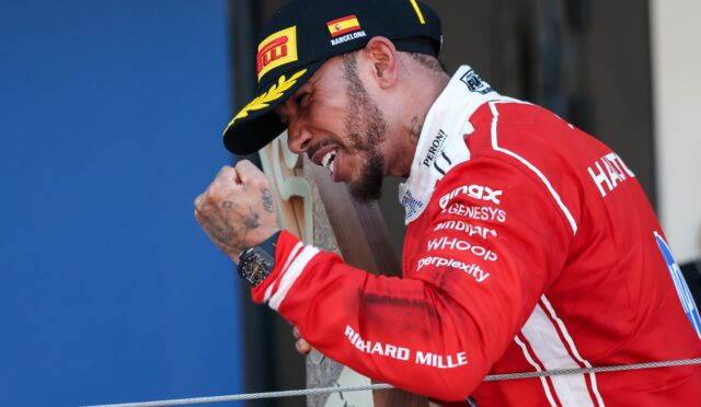 Male cyclist in a red racing suit and black cap rests his head on a wooden panel during a podium moment.