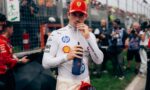 Race car driver in white suit and red Ferrari cap drinks from a blue bottle at a pit‑lane/trackside area with fans behind a fence in the background.