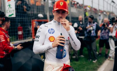 Race car driver in white suit and red Ferrari cap drinks from a blue bottle at a pit‑lane/trackside area with fans behind a fence in the background.