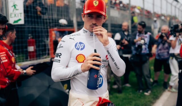 Race car driver in white suit and red Ferrari cap drinks from a blue bottle at a pit‑lane/trackside area with fans behind a fence in the background.