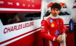 Male Formula 1 driver in a red Ferrari racing suit stands in a pit garage, holding a towel with earphones around his neck and helmets in the background.