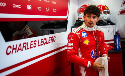 Male Formula 1 driver in a red Ferrari racing suit stands in a pit garage, holding a towel with earphones around his neck and helmets in the background.