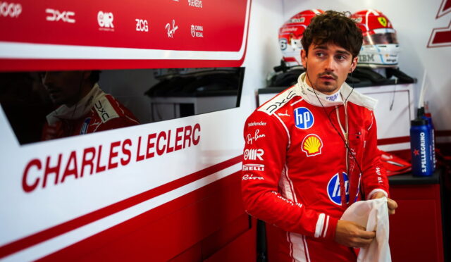 Male Formula 1 driver in a red Ferrari racing suit stands in a pit garage, holding a towel with earphones around his neck and helmets in the background.
