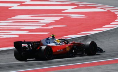 Red Formula 1 car cornering at speed on a race track, with a red-and-white curbed runoff area in the background and the driver wearing a yellow helmet.