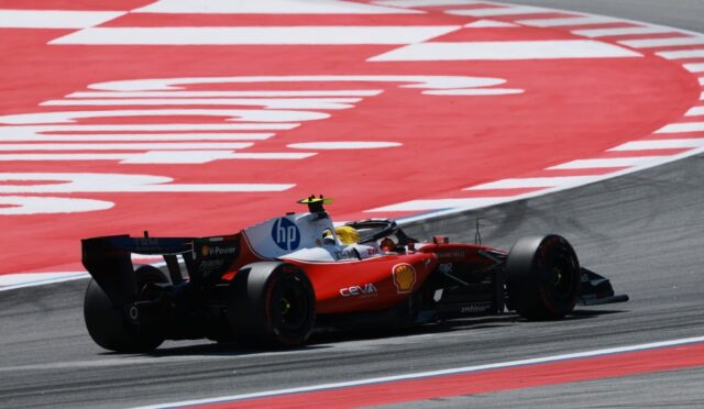 Red Formula 1 car cornering at speed on a race track, with a red-and-white curbed runoff area in the background and the driver wearing a yellow helmet.