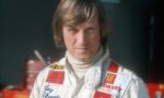 Racing driver in a white suit with red patches, standing in a garage/pit area with equipment in the background.