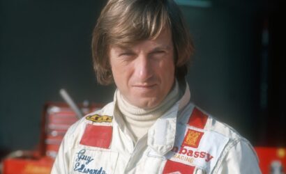Racing driver in a white suit with red patches, standing in a garage/pit area with equipment in the background.