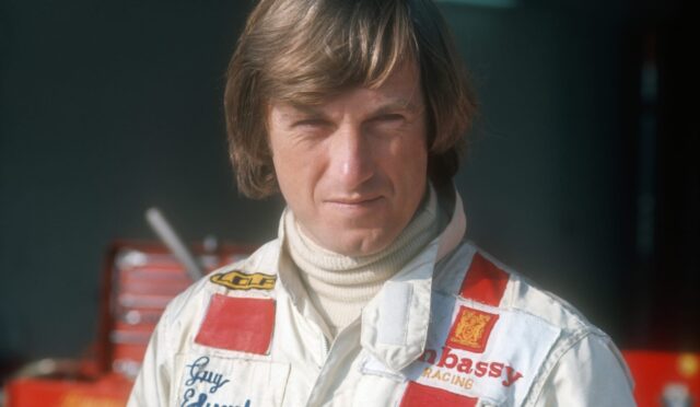 Racing driver in a white suit with red patches, standing in a garage/pit area with equipment in the background.