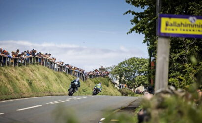 Motorcycle race on a winding rural road with two riders leaning into a bend while a crowd watches from a grassy embankment and fence.