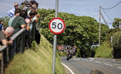 Motorcycle racer leaning into a corner on a rural road, with a crowd watching from behind a railing and a prominent 50 sign in the foreground.