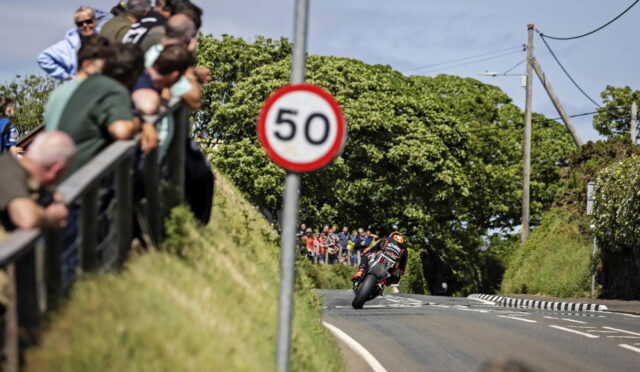 Motorcycle racer leaning into a corner on a rural road, with a crowd watching from behind a railing and a prominent 50 sign in the foreground.