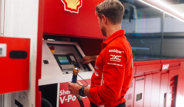 Man in a red Shell uniform uses a payment terminal at a red service kiosk indoors.