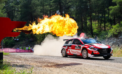 Rally car in a forest rally with a huge flame jet erupting from the exhaust, spectators behind a red barrier.
