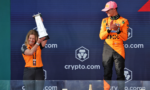 A woman on the left holds a tall trophy overhead while a man in an orange racing suit stands to the right on a podium applauding.