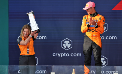 A woman on the left holds a tall trophy overhead while a man in an orange racing suit stands to the right on a podium applauding.