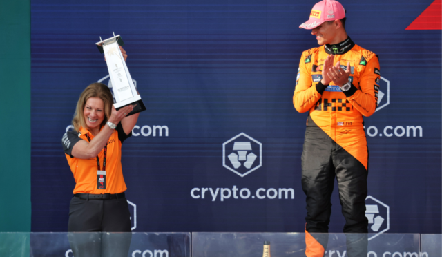 A woman on the left holds a tall trophy overhead while a man in an orange racing suit stands to the right on a podium applauding.