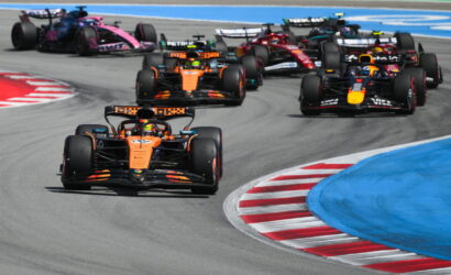 Orange Formula 1 car leads a pack around a curving track with blue runoff areas and red-and-white curbs ahead of the pack.