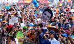 Crowd at an outdoor event taking photos; a child holds a large cutout of a smiling man’s face above the crowd.