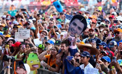 Crowd at an outdoor event taking photos; a child holds a large cutout of a smiling man’s face above the crowd.