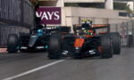Two Formula 1 cars race side by side on a city street circuit, one orange McLaren in the lead with a dark Mercedes close behind, barriers visible on the sides.