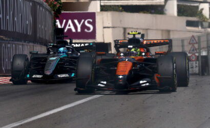 Two Formula 1 cars race side by side on a city street circuit, one orange McLaren in the lead with a dark Mercedes close behind, barriers visible on the sides.