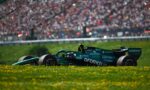 Green Formula 1 car on a race track with a cheering crowd in the blurred background and yellow flowers in the foreground.