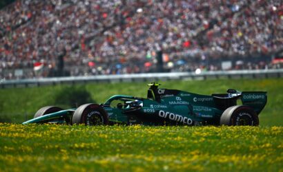 Green Formula 1 car on a race track with a cheering crowd in the blurred background and yellow flowers in the foreground.