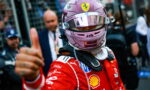Formula 1 driver in a red Ferrari racing suit and pink helmet gives a thumbs-up in the pit area.