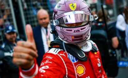 Formula 1 driver in a red Ferrari racing suit and pink helmet gives a thumbs-up in the pit area.