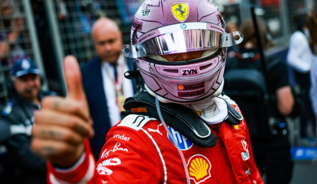 Formula 1 driver in a red Ferrari racing suit and pink helmet gives a thumbs-up in the pit area.