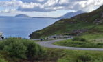 Crowd gathered along a winding coastal road on a green hillside, with the blue sea and distant islands in the background.
