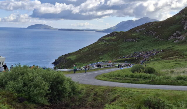 Crowd gathered along a winding coastal road on a green hillside, with the blue sea and distant islands in the background.