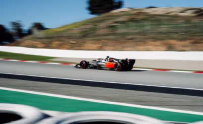 Blurred silver and black race car with orange accents speeding along a race track on a sunny day, green runoff area in the foreground.