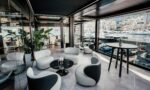 Modern harbor-side lounge with glass walls, white curved chairs, black marble tables, green plants, and a marina full of yachts outside the window.