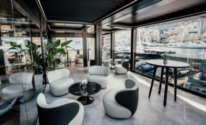 Modern harbor-side lounge with glass walls, white curved chairs, black marble tables, green plants, and a marina full of yachts outside the window.