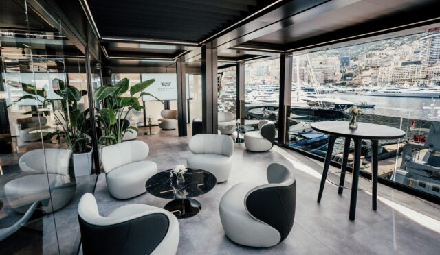 Modern harbor-side lounge with glass walls, white curved chairs, black marble tables, green plants, and a marina full of yachts outside the window.