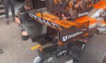 Mechanic in a wheelchair works on the rear wing of an orange and black Formula 1 car in the pit area.