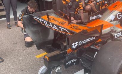 Mechanic in a wheelchair works on the rear wing of an orange and black Formula 1 car in the pit area.