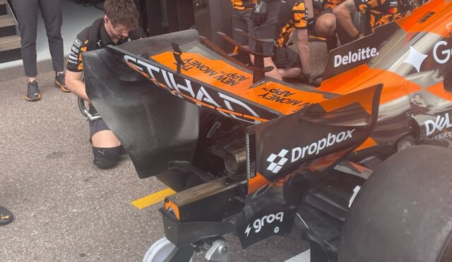 Mechanic in a wheelchair works on the rear wing of an orange and black Formula 1 car in the pit area.