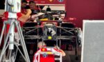 Ferrari Formula 1 race car in a garage with crew members studying blueprints or notes nearby.