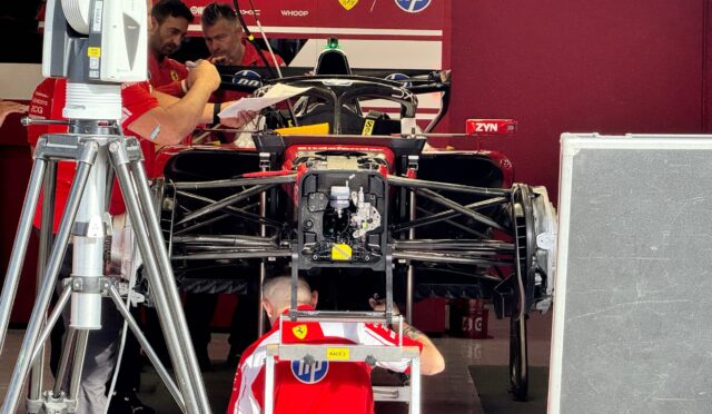 Ferrari Formula 1 race car in a garage with crew members studying blueprints or notes nearby.