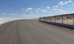 Curved race track with concrete barrier on the left and chain-link fencing on the right under a blue sky with white clouds
