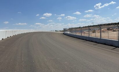 Curved race track with concrete barrier on the left and chain-link fencing on the right under a blue sky with white clouds