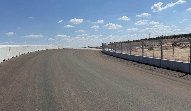 Curved race track with concrete barrier on the left and chain-link fencing on the right under a blue sky with white clouds