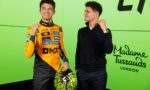 Two men pose and smile against a bright green wall; one in a racing orange suit holding a neon-yellow helmet, the other in a dark sweater with a pink watch strap visible
