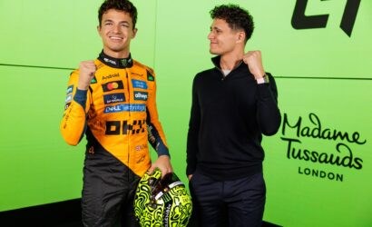 Two men pose and smile against a bright green wall; one in a racing orange suit holding a neon-yellow helmet, the other in a dark sweater with a pink watch strap visible