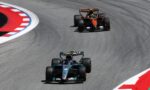 Three Formula 1 cars race through a sharp corner on a track, with red-and-white curbs along the inside edge.